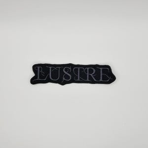 Könnte beinhalten: Ein schwarzer, gestickter Aufnäher mit dem Wort "LUSTRE" in grauem Garn. Der Aufnäher hat eine unregelmäßige Form und ist zum Aufnähen auf Kleidung oder Accessoires gedacht. Der Text ist in einer stilisierten, gotischen Schriftart gehalten.