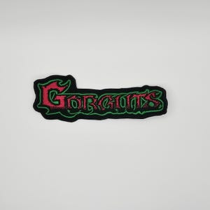 Pode incluir: Patch bordado preto com a palavra "GORGUTS" em vermelho e verde. O patch tem uma borda preta e é projetado para roupas ou acessórios.