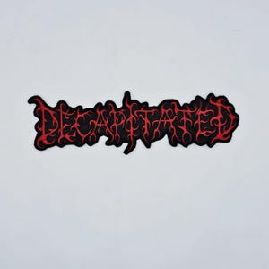 May include: A black patch with the word "DECAPITATED" in red, stylized lettering. The letters have a flame-like design, and the patch has a red border. The patch is likely for clothing or accessories.