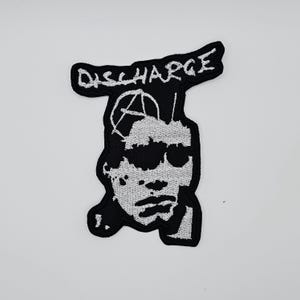 Puede incluir: Parche bordado en blanco y negro con la palabra "DISCHARGE" sobre una cara estilizada y un símbolo de anarquía. El parche está sobre un fondo blanco.