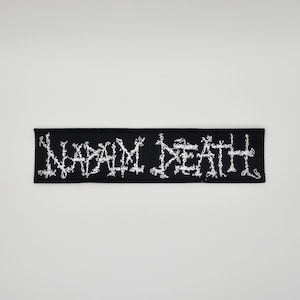 May include: Black rectangular patch with the text "NAPALM DEATH" embroidered in white. The text has a distressed, jagged font style. The patch is likely for clothing or accessories.