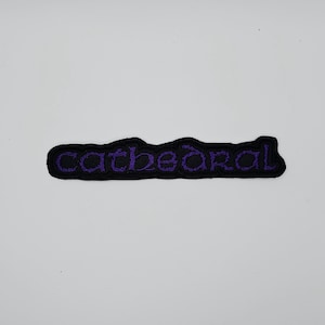 May include: A black embroidered patch featuring the word "cathedral" in purple stitching. The patch is a rounded rectangle, designed for application to clothing or other textiles.