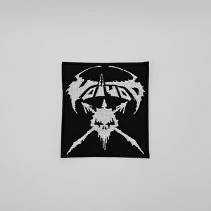 Puede incluir: Parche bordado en blanco y negro de forma cuadrada. Presenta un diseño de calavera estilizada y la palabra "VOID" en fuente gótica. El diseño blanco destaca sobre un fondo negro, ideal para ropa o accesorios.