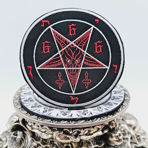 Op de afbeelding: Een zwarte en rode geborduurde patch met een pentagram, een geitenkop en het getal 666. De patch is op een zilveren standaard.