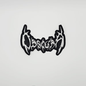 May include: A black and white embroidered patch with the word "Obsessed" in a stylized font. The patch has a black background with white stitching outlining the letters and the border. The patch is centered on a white background.