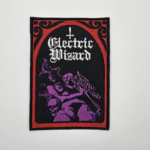 Puede incluir: Parche rectangular negro con borde rojo y las palabras "Electric Wizard" en letras góticas blancas. El parche presenta una ilustración púrpura de figuras y un esqueleto. Una cruz está encima del texto.