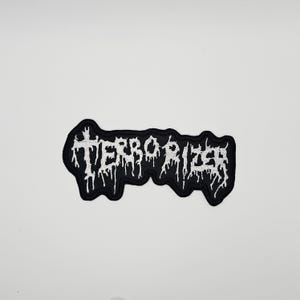 May include: Black and white embroidered patch with the word "TERRORIZER" in a gothic font. The patch has a black background with white stitching for the text and border. The patch is likely for clothing or accessories.