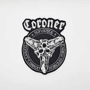 Könnte beinhalten: Ein schwarz-weiß gestickter Aufnäher mit dem Wort "Coroner" oben. Darunter befindet sich ein kreisförmiges Emblem mit einem schädelartigen Design und den Namen "Ron Royce", "Tommy Baron" und "Marquis Marky".