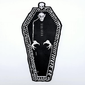 Peut inclure: Patch thermocollant brodé noir et blanc représentant un vampire dans un cercueil. Le vampire a un visage pâle et porte une robe noire. Le cercueil est décoré d'un motif squelettique.