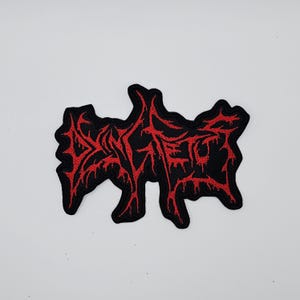 Puede incluir: Parche bordado negro con la palabra "DEATH" en rojo, con una fuente estilizada y goteante. El parche tiene una forma irregular y está diseñado para ropa o accesorios.