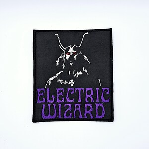 Könnte beinhalten: Schwarzer quadratischer Aufnäher mit einer weißen Illustration einer gehörnten Figur mit roten Augen. Die Worte "ELECTRIC WIZARD" sind in lila, gotischer Schrift gestickt. Der Aufnäher hat einen schwarzen Rand.
