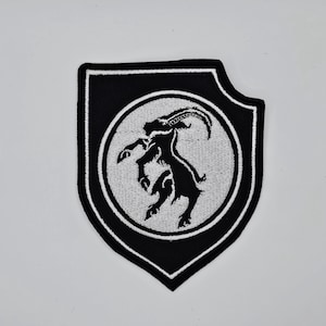 Puede incluir: Parche bordado en blanco y negro con la silueta de una cabra estilizada dentro de un círculo blanco. El parche tiene forma de escudo con un borde negro, ideal para ropa o accesorios.