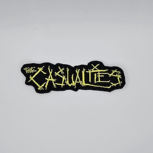 May include: Black embroidered patch featuring the text "THE CASUALTIES" in yellow. The patch has an irregular shape and is designed to be sewn or ironed onto clothing or accessories.