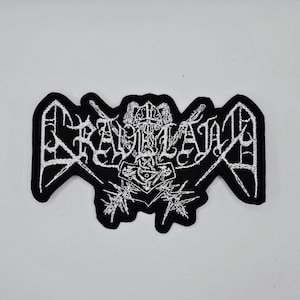 Op de afbeelding: Zwart en wit geborduurde patch met het woord "RAVELAND" in een gotisch lettertype, met een gestileerd ontwerp van vleugels en een hamer. De patch heeft een unieke vorm en is waarschijnlijk voor kleding of accessoires.