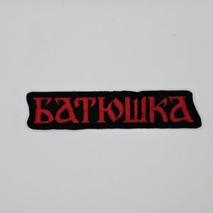 May include: Black embroidered patch with the word "Батюшка" in red, stylized Cyrillic lettering. The patch has a rectangular shape with a black border and is likely designed to be sewn or ironed onto fabric.