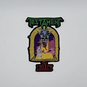 Puede incluir: Parche bordado con el nombre de la banda "TESTAMENT" en verde. El diseño presenta una calavera con cuernos, una llama y el texto "THE LEGACY" en rojo, todo dentro de un marco gótico.