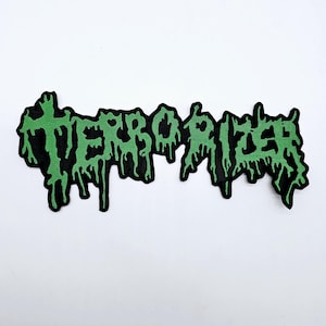 May include: A black patch with the word "TERRORIZER" in dripping green letters. The patch has a black border and is set against a white background. The design has a distressed, grunge aesthetic.