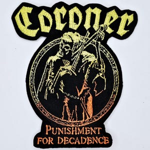 Peut inclure: Écusson noir brodé avec le mot "Coroner" en lettres gothiques jaunes. L'écusson représente une figure squelettique jouant d'un instrument à cordes, avec les mots "Punishment for Decadence" en dessous.