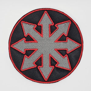 Op de afbeelding: Ronde zwarte patch met een grijs achtpuntig sterontwerp, waarbij elke punt een pijl is. De ster is rood omlijnd en de patch heeft een rode rand. Het is waarschijnlijk een geborduurd embleem.