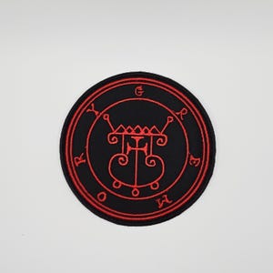 Könnte beinhalten: Ein runder, schwarzer, gestickter Aufnäher mit einem roten Sigill-Design. Das Sigill weist komplizierte Linien, Symbole und Buchstaben innerhalb eines kreisförmigen Randes auf. Der Aufnäher ist für Kleidung oder Accessoires geeignet.