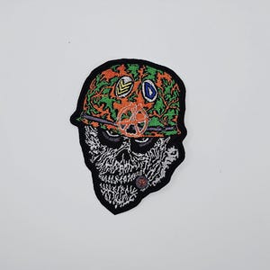 May include: Embroidered patch featuring a skull wearing a camouflage helmet with orange, green, and yellow accents. The skull has a lit cigar and a peace sign. The patch has a black border.