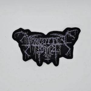 May include: Black embroidered patch with the text "Forgotten Tomb" in a gothic font. The patch has a unique, irregular shape with a black border and light gray lettering. The design has a distressed, vintage aesthetic.