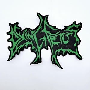 Puede incluir: Un parche negro con un diseño verde estilizado. El diseño verde parece ser una palabra o frase en una fuente única. El parche tiene una forma irregular y dentada, y probablemente sea un parche para planchar o coser.