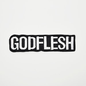 May include: A black and white embroidered patch with the word "GODFLESH" in bold, white capital letters. The patch has a black border and is rectangular in shape. The patch is centered on a white background.