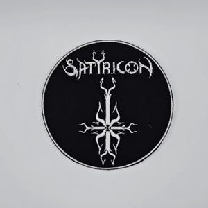 May include: A circular black patch with a white embroidered design. The design features the word "SATYRICON" in a stylized font above a white cross-like symbol. The patch has a white border.