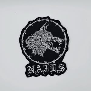 Pode incluir: Patch bordado a preto e branco com a imagem de uma cabeça de lobo rosnando dentro de um círculo de arame farpado. A palavra "NAILS" é bordada em letras góticas brancas por baixo. Para roupa ou acessórios.