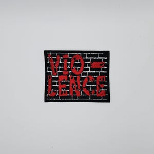 Könnte beinhalten: Ein schwarzer, rechteckiger Aufnäher mit dem Wort "VIOLENCE" in roter, stilisierter Schrift. Der Text ist über einem Backsteinmauer-Design mit schwarzen Ziegeln und weißem Mörtel angeordnet. Für Kleidung oder Accessoires.