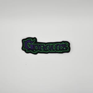 May include: Black embroidered patch with the word "Gorgons" in a stylized font. The text is in purple with a green outline, set against a black background. The patch has a decorative border.