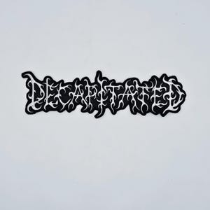 May include: Black and white embroidered patch with the word "DECAPITATED" in a stylized font. The text is white, outlined in black, and set against a black background. The patch has a unique, irregular shape.