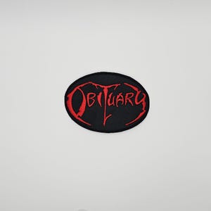 Peut inclure: Écusson ovale noir avec le mot "Obituary" brodé en rouge dans une police stylisée. Le patch a une bordure noire et est posé sur un fond blanc. Le texte est dans une police gothique.