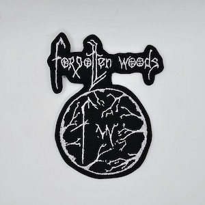 May include: Black and white embroidered patch featuring the text "forgotten woods". The patch has a circular design with a tree-like pattern. This patch is designed to be sewn onto clothing or accessories.