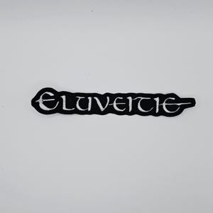 Puede incluir: Parche bordado en blanco y negro con la palabra "ELUVEITIE" en una fuente estilizada. El parche tiene un borde negro y está diseñado para ser cosido o planchado sobre tela.