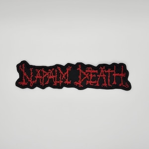 Puede incluir: Un parche bordado negro con la inscripción "NAPALM DEATH" en letras rojas. El texto es estilizado y el contorno del parche es negro. El diseño evoca el estilo del metal o punk.