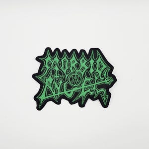 Könnte beinhalten: Ein schwarzer Aufnäher mit grün gesticktem Text und Symbolen. Der Aufnäher hat einen gezackten Rand und zeigt ein stilisiertes Design mit einem Pentagramm. Die grüne Stickerei hebt sich vom schwarzen Hintergrund ab.
