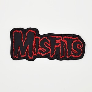 Peut inclure: Écusson noir avec le mot "MISFITS" brodé en rouge, bordé d'un contour rouge. L'écusson a une forme irrégulière unique et est posé sur un fond blanc.