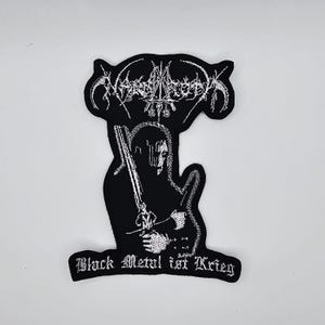 May include: Black embroidered patch with a white border, featuring a figure with a sword and the text "Black Metal ist Krieg". The patch has a black background and a stylized design at the top.