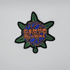 May include: Embroidered patch featuring a blue and orange floral design with the word "SLEEP" in orange. The patch has a black border with green leaf accents. The patch is likely for clothing or accessories.