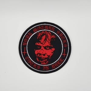 Peut inclure: Écusson rond noir avec un visage démoniaque rouge au centre. Le texte "YOUR MOTHER SUCKS COCKS IN HELL" est brodé en rouge autour du visage. Une bordure blanche entoure l'écusson noir.