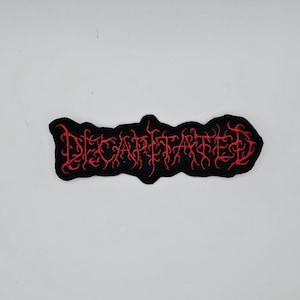 Puede incluir: Parche bordado negro con la palabra "DECAPITATED" en rojo, con letras góticas. El parche tiene una forma irregular y está diseñado para ser cosido en ropa o accesorios. El texto está en una fuente gótica.