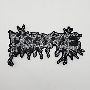 May include: Black patch with white embroidered text. The text appears to be a stylized word, possibly a band name or logo, with a distressed, organic design. The patch has a jagged, irregular border.