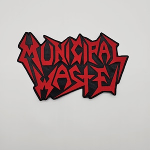 Puede incluir: Un parche bordado en negro y rojo con las palabras "MUNICIPAL WASTE" en una fuente estilizada y dentada. El texto es rojo sobre un fondo negro. El parche está diseñado para coser en ropa o bolsos.