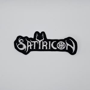 Könnte beinhalten: Schwarz-weißer gestickter Aufnäher mit dem Wort "SATYRICON" in einer stilisierten Schriftart. Der Aufnäher zeigt ein Dreizack-Symbol über dem Buchstaben "A" und ein kreuzartiges Emblem auf der rechten Seite. Der Aufnäher befindet sich auf weißem Hintergrund.