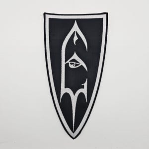 Könnte beinhalten: Schwarzer schildförmiger Aufnäher mit weißem Stickdesign. Das Design zeigt ein stilisiertes Auge und eine buchstabenartige Form. Der Aufnäher hat einen weißen Rand und ist für Kleidung oder Accessoires geeignet.