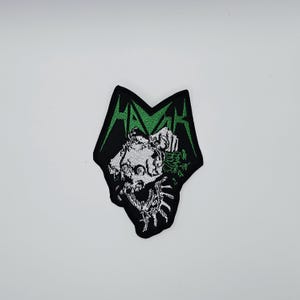 Op de afbeelding: Geborduurde patch met een zwart en groen ontwerp. De patch toont het woord "HAMR" in groene, gestileerde letters. Onder de tekst staat een gedetailleerde illustratie van een schedel met een hand en groen gebladerte. De patch heeft een unieke, hoekige vorm.