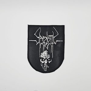 Może przedstawiać: Czarna naszywka w kształcie tarczy z białym haftowanym tekstem i grafiką demonicznej twarzy. Tekst brzmi "Sadistic Intent" w gotyckiej czcionce. Idealna do naszywania na odzież lub torby.