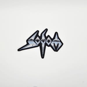May include: Embroidered patch featuring the word "Sodom" in a stylized, angular font. The patch has a black border with light blue and white detailing within the letters. The patch is likely for clothing or accessories.
